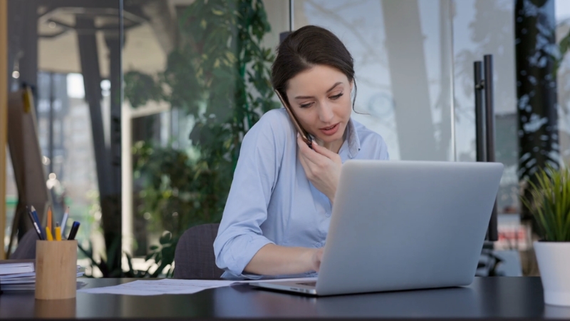 Žena razgovara telefonom i istovremeno radi na laptopu za stolom kod kuće