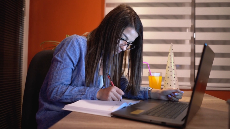 Mlada žena sedi za radnim stolom, koristi laptop i zapisuje beleške u svesku